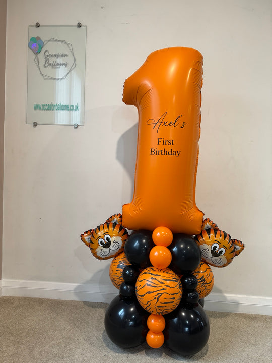 A custom first birthday balloon decoration featuring a large inflatable orange number 1 balloon with the text 'Ariel's First Birthday', supported by a cluster of tiger-themed balloons and black and orange latex balloons on a carpeted floor against a plain wall.