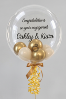 A large clear round balloon with black script text reading 'Congratulations on your engagement Oakley & Kiara,' filled with metallic gold and cream latex balloons, tied with a gold ribbon and hanging gold confetti on a white background.