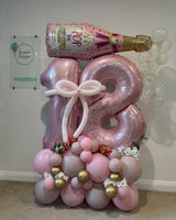 Pink 18 number balloons with gold personalisation, a white balloon bow, champagne bottle and clear balloon bubbles above a floral pink and gold base.