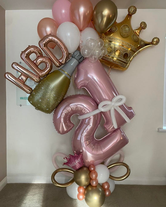  Pink 21 foil balloons with a white bow, gold crown, champagne bottle, rose gold HBD lettering and coordinating pink, white and gold balloons.