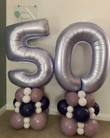 Two large metallic silver number 50 foil balloons standing on coordinated balloon arrangements of dusty lavender, white, and dark navy, photographed indoors against a plain wall.