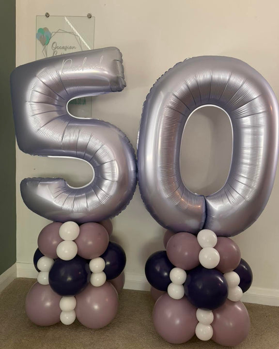 Two large metallic silver number 50 foil balloons standing on coordinated balloon arrangements of dusty lavender, white, and dark navy, photographed indoors against a plain wall.