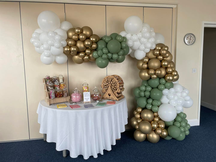 A decorated event setup with a white draped table holding jars of candy and small items, set against a balloon arch in white, gold, and sage green inside a room with beige paneled walls.