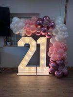 A large illuminated marquee number 21 on a wooden floor, decorated with pink, rose gold, and white balloon garlands in a room setting.