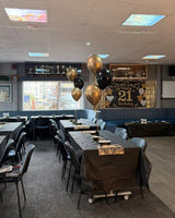 A large indoor event space or banquet hall with multiple tables covered in dark tablecloths, chairs, a ceiling-mounted projector, black and gold balloons, and a decorative '21' sign on the wall. A person is visible near a suitcase in the background.