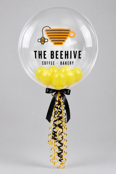 Clear bubble balloon printed with The Beehive Coffee Bakery logo, filled with yellow balloons and finished with black and yellow ribbons.