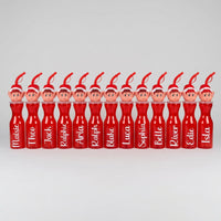 A row of personalized red plastic drink bottles with elf character heads on top, each printed with a different name, set against a plain indoor wall and carpeted floor.