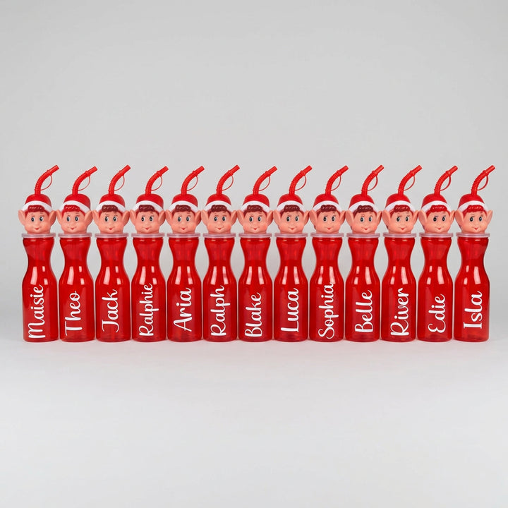 A row of personalized red plastic drink bottles with elf character heads on top, each printed with a different name, set against a plain indoor wall and carpeted floor.