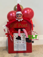 Red Elf Post box topped with red and gold balloons, featuring a seated Christmas elf, candy cane, red bow and elf surveillance sign.