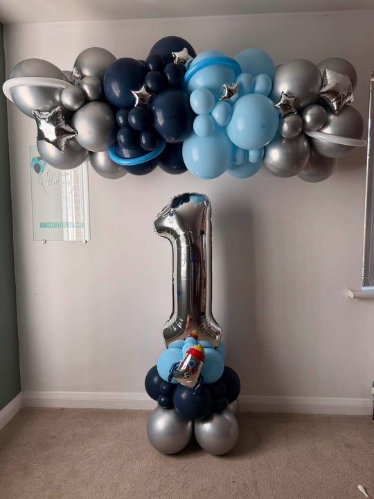 Silver number 1 balloon with a rocket on a blue and silver base, beneath a navy, light blue and silver garland with stars and balloon planet rings.