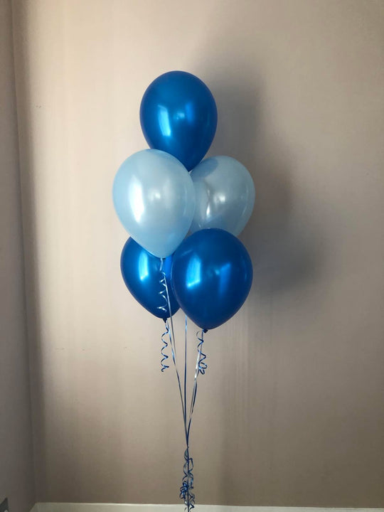 A cluster of six metallic balloons in shades of royal blue and light ice blue, tied together with blue ribbons against a plain beige wall.