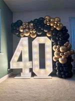 Large illuminated marquee numbers '40' on a carpeted floor, decorated with a black and gold balloon arch behind them in an indoor room.