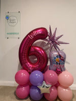 A birthday balloon arrangement featuring a large metallic pink number 6 foil balloon with the name 'Willen' written on it, surrounded by pink, purple, and light pink latex balloons, a purple star balloon, and a heart-shaped balloon with text, set against a plain indoor wall.