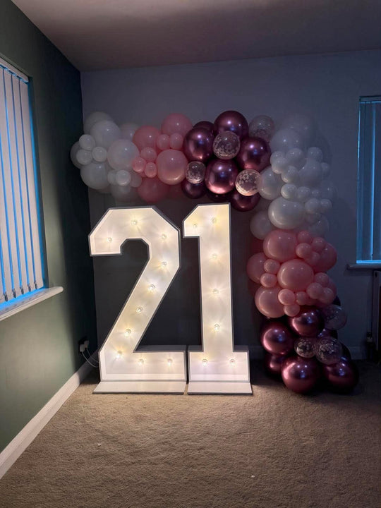 Large illuminated '21' letter numbers with built-in lights, styled in a room with a balloon garland backdrop in pink, white, and metallic rose gold.