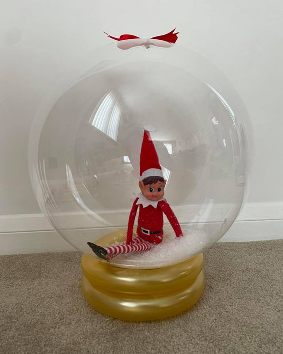 Clear elf arrival snow globe balloon containing a red Christmas elf and artificial snow, finished with a gold balloon base and red and white bow.
